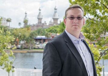 A person in a suit stands outdoors near a river with historic buildings and trees in the background.